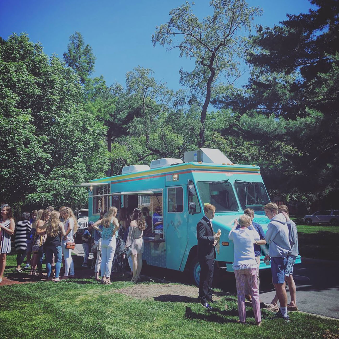 People standing in line at a bright blue food truck parked in a sunny, tree-filled park. People standing in line at a bright blue food truck parked in a sunny, tree-filled park.